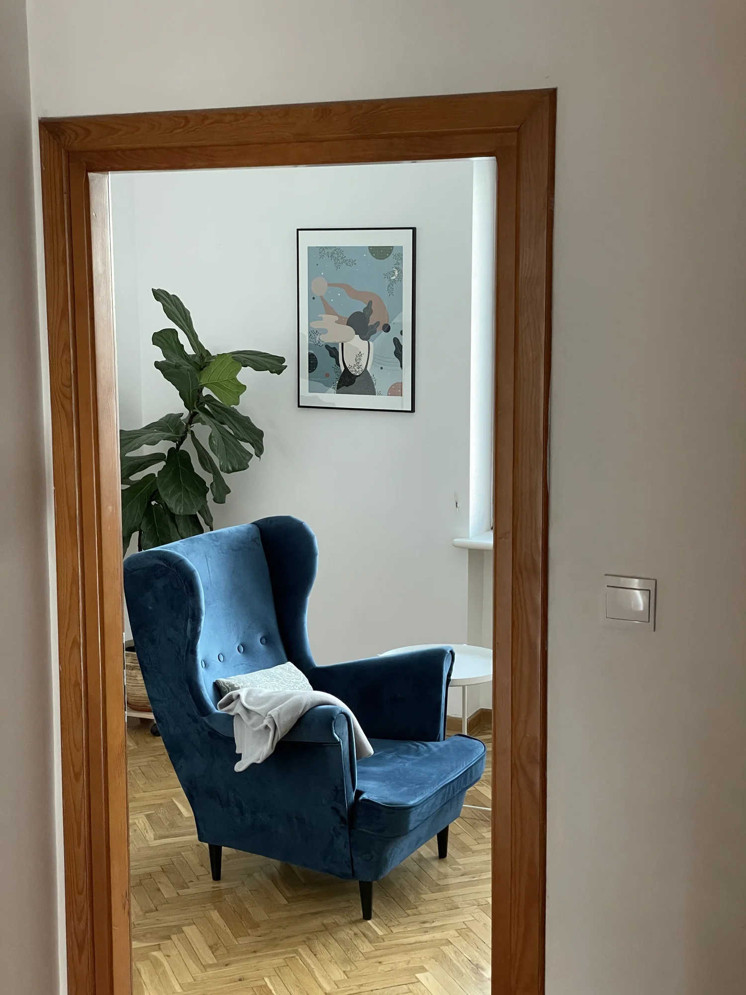 Gabinet psychoterapii na Saskiej Kępie widziany przez otwarte drzwi – niebieski fotel uszak z kocem, fikus i grafika na ścianie